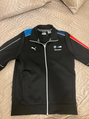 Svart BMW Motorsport jacka från Puma - Svart jacka från Puma x BMW Motorsport med dragkedja, ståkrage och loggor på bröstet. Snygga detaljer i blått och rött på axlarna samt vita piping längs ärmarna. Perfekt för dig som gillar motorsport och streetwear.