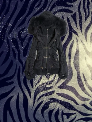 Svart parkajacka med fuskpäls - Säljer en svart parkajacka med stor huva och fuskpälsdetalj. Jackan har dragkedja, snygga spännen framtill och ribbade muddar vid ärmslut. Perfekt för kalla dagar och har en riktigt chill streetstil.