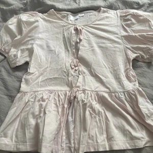 Ljusrosa blus med knyt och puffärm - Supersöt ljusrosa blus med puffärmar och tre knyt framtill. Blusen har en volang nedtill och är tillverkad i mjuk bomull. Perfekt för dig som gillar romantiska detaljer och en luftig känsla.