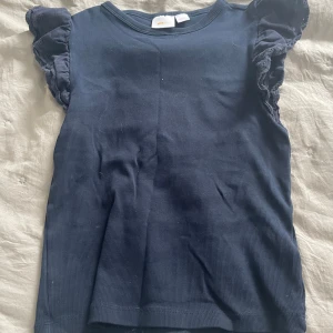 Mörkblå ribbad topp med volangärm - Säljer en mörkblå ribbad topp från Gina Tricot med korta volangärmar. Toppen har rund hals och är tillverkad i mjuk bomull. Perfekt för dig som gillar en enkel men söt stil med extra detaljer på ärmarna.