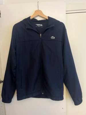 Mörkblå vindjacka från Lacoste - Säljer en mörkblå vindjacka från Lacoste med huva och dragkedja framtill. Pris kan diskuteras vid snabb affär
