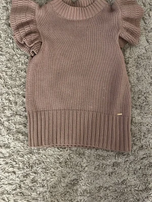 Beige stickad topp Holly & Whyte - Säljer en beige stickad topp från Holly & Whyte by Lindex i storlek XS. Toppen har ribbad hög hals, bred ribbad kant nedtill och korta ärmar med volangdetalj. Perfekt för dig som gillar en söt och trendig look.💞💞