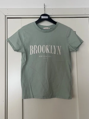 Grön t-shirt Brooklyn KappAhl - Snygg ljusgrön t-shirt från KappAhl med trycket 'BROOKLYN NEW YORK CITY' på bröstet. Klassisk rund halsringning och korta ärmar. Skön och mjuk i bomull, perfekt till en chill vardagslook.