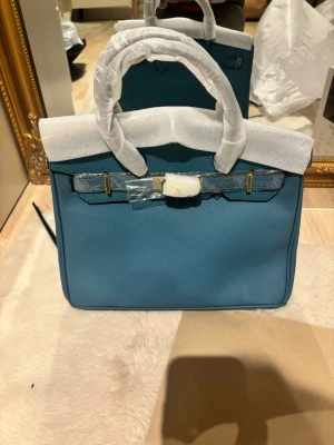 Blå handväska i skinn med gulddetaljer - Säljer en stilren blå handväska i skinn med guldfärgade metalldetaljer och klassisk form. Väskan har dubbla handtag och strukturerad yta. Perfekt för dig som vill ha en snygg accessoar med exklusiv känsla.