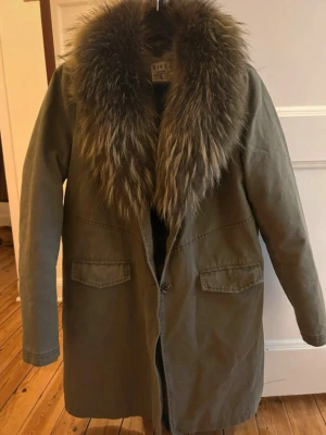 Olivgrön parkas - Säljer en olivgrön parkas från FIVE  med stor, fluffig pals runt kragen. Jackan har två stora fickor framtill, knappstängning och rak passform. Perfekt för kalla dagar och ger en riktigt snygg streetstyle-vibe. Nypris 4000kr