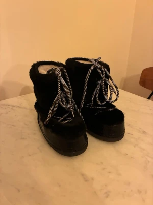 Svarta moon boots från Guess - Svarta moon boots från Guess med fluffig pälsimitation och breda, svartvita snören. Skorna har en chunky platt sula och högre skaft som håller värmen. Perfekta för vintern och riktigt snygga till streetwear. Guess-logga baktill.