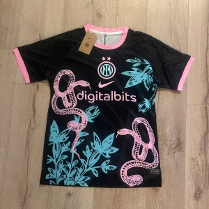 Inter Milan x Nike svart fotbollströja - Säljer en Inter Milan fotbollströja från Nike i svart med rosa och turkosa detaljer. Tröjan har korta ärmar, rosa ormmotiv och turkosa växtmönster framtill, samt rosa kant vid hals och ärmslut. Materialet är lätt och andas, perfekt för match eller träning.