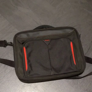 Svart datorväska från Targus - Praktisk svart datorväska från Targus med röda detaljer på framsidan. Väskan har flera fack för mobil, pennor och tillbehör, samt en justerbar axelrem. Insidan är organiserad med tydliga fack och elastiska band för USB och dokument. Katt finns i hemmet