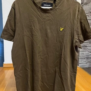 Brun t-shirt från Lyle & Scott - Snygg brun t-shirt från Lyle & Scott med klassisk rund hals och korta ärmar. På bröstet finns den ikoniska gula fågeln broderad. T-shirten är gjord i mjuk bomull och har en enkel, stilren design som funkar till det mesta.