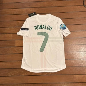 Portugal Ronaldo EM 2012 fotbollströja - Säljer en officiell Portugal fotbollströja från EM 2012 med Ronaldo och nummer 7 på ryggen. Tröjan är vit med gröna och röda korsande ränder, korta ärmar och Euro 2012-patch på båda ärmarna. Tillverkad i lätt Dri-Fit material som andas.