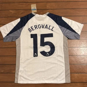Tottenham Bergvall #15 Nike tröja M - Snygg Tottenham Hotspur fotbollströja med Bergvall och nummer 15 på ryggen. Tröjan är vit med marinblå och grå detaljer, har korta ärmar och är tillverkad i ventilerande polyester. Framsidan har Spurs-logga, Nike Swoosh och AIA i rött. Perfekt för dig som älskar Premier League.
