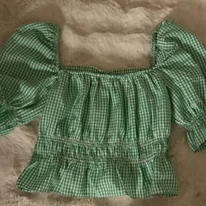 Grön rutig blus med puffärmar - Säljer en söt grön blus med vitt rutmönster och puffiga korta ärmar. Toppen har smockad midja och volang nedtill, samt offshoulder-design som ger en trendig vibe. Perfekt för dig som gillar romantisk och lekfull stil.
