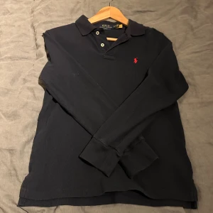 Svart långärmad pikétröja Polo Ralph Lauren - Klassisk svart långärmad pikétröja från Polo Ralph Lauren i slim fit-modell. Tröjan har krage, knäppning med två knappar och den ikoniska röda broderade loggan på bröstet. Perfekt för en stilren och tidlös look. Passar dig som är ungefär 165