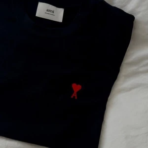 Svart t-shirt från Ami med rött hjärta - Svart t-shirt från Ami Alexandre Mattiussi med klassisk passform och rund hals. På bröstet finns Ami:s ikoniska röda hjärta med bokstaven A broderat. T-shirten är i mjuk bomull och har korta ärmar. Perfekt för en clean och stilren look. Försten att betala får Varan. Priset är satt 