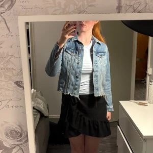 Croppad jeansjacka med rå kant - Säljer en croppad jeansjacka i ljusblå denim med rå, fransad nederkant. Jackan har klassiska bröstfickor med knappar och silverfärgade detaljer. Perfekt för dig som gillar en chill och trendig stil.