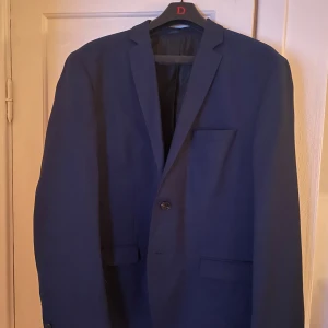 Mörkblå kavaj Jack & Jones strl 54 och byxor - Snygg mörkblå kavaj från Jack & Jones Premium i klassisk modell med två framfickor och bröstficka. Kavajen har enkelknäppning och stilren design, perfekt för dig som gillar en clean look. Materialet är mjukt och känns bekvämt mot kroppen. Det medföljer även ett par matchande kostymbyxor. (ANVÄND EN GÅNG)