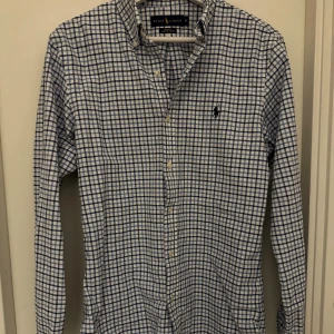 Rutig blåvit skjorta från Ralph Lauren - Snygg slim fit skjorta från Ralph Lauren i stretchig bomull. Skjortan är blåvit med rutigt mönster, har klassisk krage, knappar framtill och lång ärm. Ikoniska Polo-loggan broderad på bröstet. Perfekt för dig som gillar stilren och tidlös design.