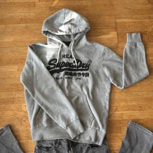 Grå hoodie från Superdry - Säljer en grå hoodie från Superdry med svart logga och japansk text på bröstet. Tröjan har huva med snörning, känguruficka och långa ärmar. Perfekt för dig som gillar en avslappnad och sportig stil.