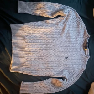 Grå kabelstickad tröja Polo Ralph Lauren - Kabelstickad grå tröja från Polo Ralph Lauren med rund halsringning och broderad logga på bröstet. Tröjan är i mjuk ull och har långa ärmar samt ribbade muddar. Perfekt för lager-på-lager under kyliga dagar. Storleken är L
