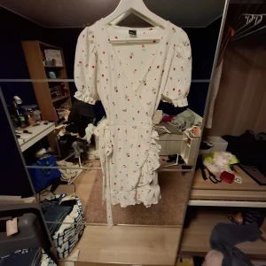 Vit blommig omlottklänning Gina Tricot - Supersöt vit omlottklänning från Gina Tricot med små röda blommor och gröna blad. Klänningen har puffiga korta ärmar med volangkant, v-ringning och knytband i midjan. Materialet är mjukt och lätt, perfekt för varmare dagar. Volangdetaljer längs nederkanten ger extra charm. Aldrig använd bara testad tyckte dock inte att den satt bra på mig