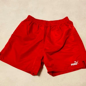 Retro Röda träningsshorts från Puma - Snygga röda shorts från Puma med elastisk midja och vit logga på benet. Tillverkade i lätt polyester som andas, perfekta för sport eller chill. Klassisk loose fit och praktiska fickor både fram och bak. Enkelt att matcha med din favorit-t-shirt.