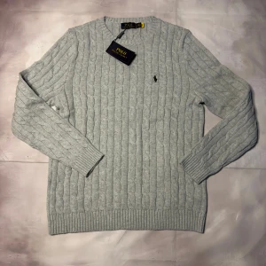 Ljusgrå stickad tröja Polo Ralph Lauren - Snygg ljusgrå stickad tröja från Polo Ralph Lauren i 100% bomull. Tröjan har klassiskt kabelstickat mönster, rund hals och ribbade muddar. Diskret broderad logga på bröstet ger en clean och stilren look. Perfekt för dig som gillar tidlös stil.