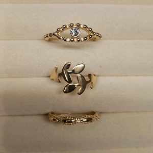 Set med tre guldiga ringar - Säljer ett set med tre snygga guldiga ringar. En ring har ett ögonformat mönster med en klar sten i mitten, en har ett bladliknande motiv och den tredje har ett tvinnat, detaljerat mönster. Perfekta för att mixa och matcha till din stil.
