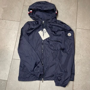 Mörkblå vindjacka  - Snygg mörkblå vindjacka med huva och coola snörningsdetaljer. (Storlek M sitter som S) Jackan har dragkedja framtill, -logga på ärmen och diskreta rödvita ränder på huvan. Perfekt för dig som gillar stilren design och exklusiva märken.