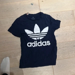 Mörkblå Adidas t-shirt med logga - Snygg mörkblå t-shirt från Adidas med stor vit trefoil-logga och text på framsidan. Klassisk rund hals och korta ärmar. Tillverkad i mjuk bomull som är skön mot huden. Perfekt för dig som gillar sportiga och stilrena plagg.