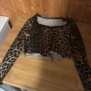 Leopardmönstrad croppad bolerotopp - Säljer en croppad bolerotopp med leopardmönster i bruna och beige toner. Toppen har långa ärmar och en rundad halsringning med svart spetskant. Perfekt för dig som vill sticka ut med ett djuriskt print och edgy vibe.