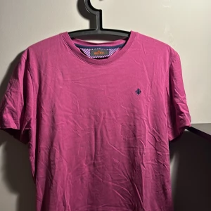 Rosa t-shirt från Morris - Snygg rosa t-shirt från Morris med rund hals och korta ärmar. T-shirten har ett litet broderat emblem på bröstet och insidan av kragen har ett lila tyg med vita prickar. Materialet är mjuk bomull, perfekt för en chill stil.