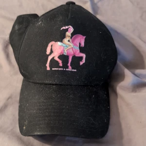 Svart keps med frökensnusk-tryck - Svart keps med coolt tryck av fröken snusn som rider på en rosa häst. Perfekt för dig som vill sticka ut med en unik och färgglad detalj. Kepsen har klassisk form och böjd skärm, tillverkad i bomull för skön passform.