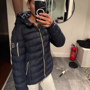 Mörkblå pufferjacka från Vinson Polo Club - Säljer en mörkblå pufferjacka från Vinson Polo Club med huva och snygga guldfärgade dragkedjor. Jackan har quiltad design, logga på ärmen och praktiska fickor. Perfekt för kyliga dagar och riktigt skön att bära.