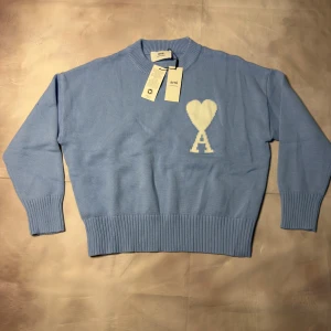 Ljusblå stickad tröja från Ami - Säljer en ljusblå stickad tröja från Ami Alexandre Mattiussi med vit hjärtformad A-logga på bröstet. Tröjan har rund hals, ribbade muddar och är tillverkad i mjuk bomull. Perfekt för dig som gillar stilrena detaljer och vill ha en chill look.