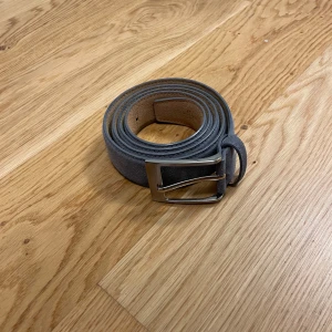Grå mockabälte med silverfärgat spänne - Snyggt grått bälte i mocka med ett stilrent, silverfärgat metallspänne. Bältet har en klassisk, rak form och passar perfekt till jeans eller chinos. Den mjuka mockan ger en cool och modern känsla till din outfit.