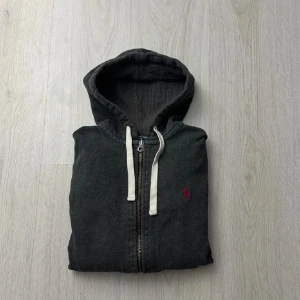 Mörkgrå hoodie från Polo Ralph Lauren - Snygg mörkgrå hoodie från Polo Ralph Lauren med dragkedja och vit snörning i huvan. Tröjan har ett rött broderat Polo logo på bröstet.enkel att matcha med jeans eller joggers. Perfekt till dig som vill ha en snygg samt varm hoodie nu till vintern 
