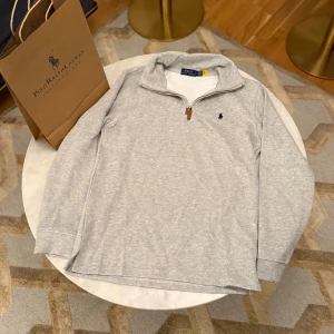 Grå half zip tröja Polo Ralph Lauren - Snygg ljusgrå långärmad tröja från Polo Ralph Lauren med klassisk broderad logga på bröstet. Tröjan har en stilren half zip med läderdetalj på dragkedjan och en mjuk bomullskänsla. Perfekt för chill dagar och enkel att matcha med jeans eller joggers.