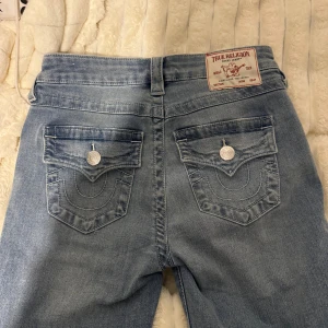 True Religion Becca Bootcut Jeans ljusblå - Ljusblå jeans från True Religion, modell Becca Mid Rise Bootcut. Klassisk femficksdesign med snygga detaljerade bakfickor och silverfärgade knappar. Jeansen har bootcut-ben och är tillverkade i mjukt denimtyg för en skön passform.