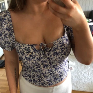 Blommig croppad T-shirt/blus  - Supersöt blommig blus i vitt och blått. Toppen har korta lite puffärmar, fyrkantig ringning och knytning framtill. ”ribbad” stretchigt rygg i bak på ryggen. Den är S/M