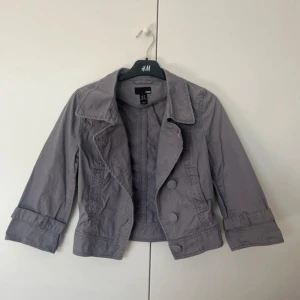 Grå trekvarts jacka  - Snygg grå kavajjacka från H&M i storlek 38. Jackan har klassisk krage, stora knappar framtill och detaljer vid ärmsluten. Perfekt för dig som gillar stilrena och enkla plagg med lite edge. Passar till många olika outfits.