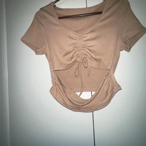Beige croppad topp med snörning - Säljer en beige croppad topp med korta ärmar och snygg snörning framtill. Toppen har ribbad struktur, v-ringning och cool cut-out detalj vid magen. Perfekt för dig som gillar trendiga och unika plagg!