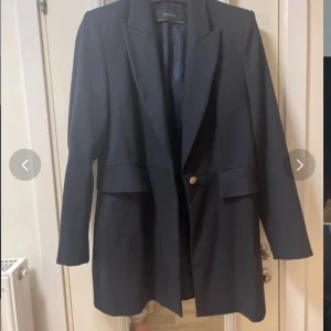 Svart kappa  från Zara Basic med guldknappar - Stilren svart kavaj från Zara Basic med snygga guldknappar på ärmslut och framtill. Kavajen har klassisk krage, långa ärmar och två fickor med lock. Perfekt för dig som vill ha en tidlös och elegant look.