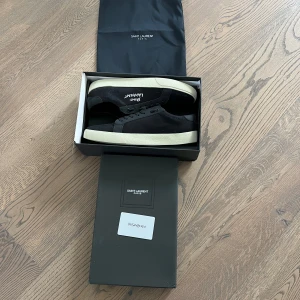 Svarta sneakers från Saint Laurent - Snygga svarta sneakers från Saint Laurent med vit sula och broderad logga på sidan. Skorna har snörning, rund tå och är tillverkade i en mix av canvas och skinn. Klassisk och stilren design med diskreta detaljer och guldtext på plösen.