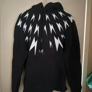 Neil Barrett - Svart hoodie från Neil Barrett med ikoniskt vitt blixtmönster över axlar och bröst. Perfekt för dig som vill sticka ut med en cool streetstyle-look, har dock en liten reva på en av blixtarna som knappt syns Stl L Skick 9/10