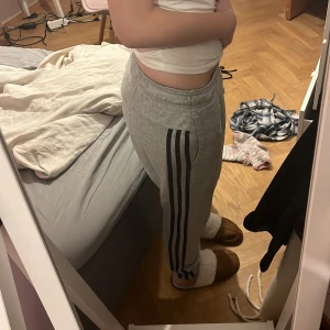 Grå Adidas mjukisbyxor med svarta ränder - Grå mjukisbyxor från Adidas med klassiska svarta ränder längs sidorna. Byxorna har resår i midjan och är tillverkade i mjukt bomullsmaterial. Perfekta för chill eller träning, med en avslappnad passform och sportig look.