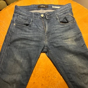 Replay Anbass blå jeans herr - Snygga blå Replay Anbass jeans med klassisk femficksdesign och diskreta slitningar. Jeansen har normal passform, raka ben och är tillverkade i mjukt denimtyg. Märkesdetaljer syns på fickorna och bak med Replay-logga. Perfekta för en avslappnad och stilren look.