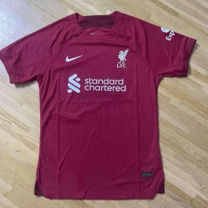 Liverpool FC matchtröja Nike röd - Säljer en officiell Liverpool FC matchtröja från Nike i klassiskt röd färg med vita loggor och sponsortryck. Tröjan har korta ärmar, rund hals och är tillverkad i lätt, ventilerande material perfekt för fotboll eller supporterliv.