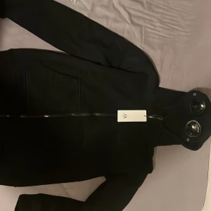 Svart gogglehoodie från C.P. Company - Storleken är Medium men är liten så passar bättre om du är S eller mellan där. Svart gogglejacka från C.P. Company med ikoniska glasögon i huvan, broderad logga på bröstet och två stora fickor framtill. Jackan har hel dragkedja och är tillverkad i mjuk polyester som ger en clean och modern look. Helt ny äkta men säljer eftersom den var för liten för mig