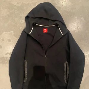 Svart Nike tech fleece  - Svart hoodie från Nike i storlek 158-170 cm med hel dragkedja framtill, två fickor med svarta detaljer och diskret Nike-logga på bröstet. Tröjan har huva och är långärmad, perfekt för en sportig och avslappnad stil.