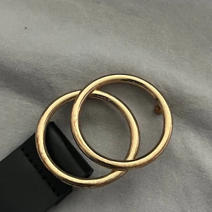 Svart bälte med guldfärgat spänne Gina Tricot - Stilrent svart bälte från Gina Tricot med ett snyggt, rundat dubbelt guldfärgat metallspänne. Bältet är slätt och har en klassisk form som passar till jeans eller kjol. Materialet är syntet och spännet ger en trendig touch till din outfit.